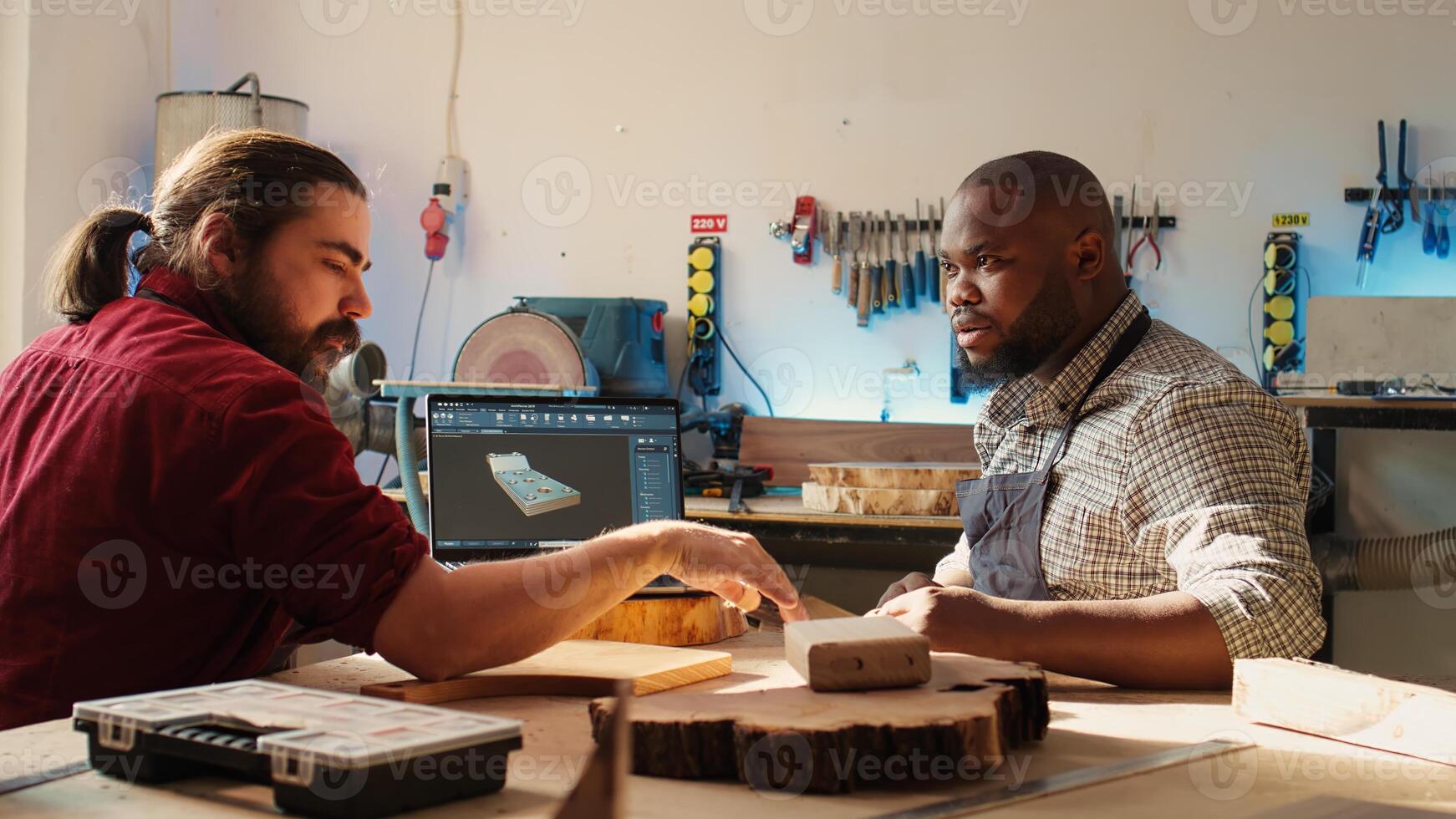 Happy BIPOC cabinetmaker and colleague comparing wood piece with schematic displayed on laptop. Cheerful carpenters in joinery crosschecking wooden object with notebook blueprint photo