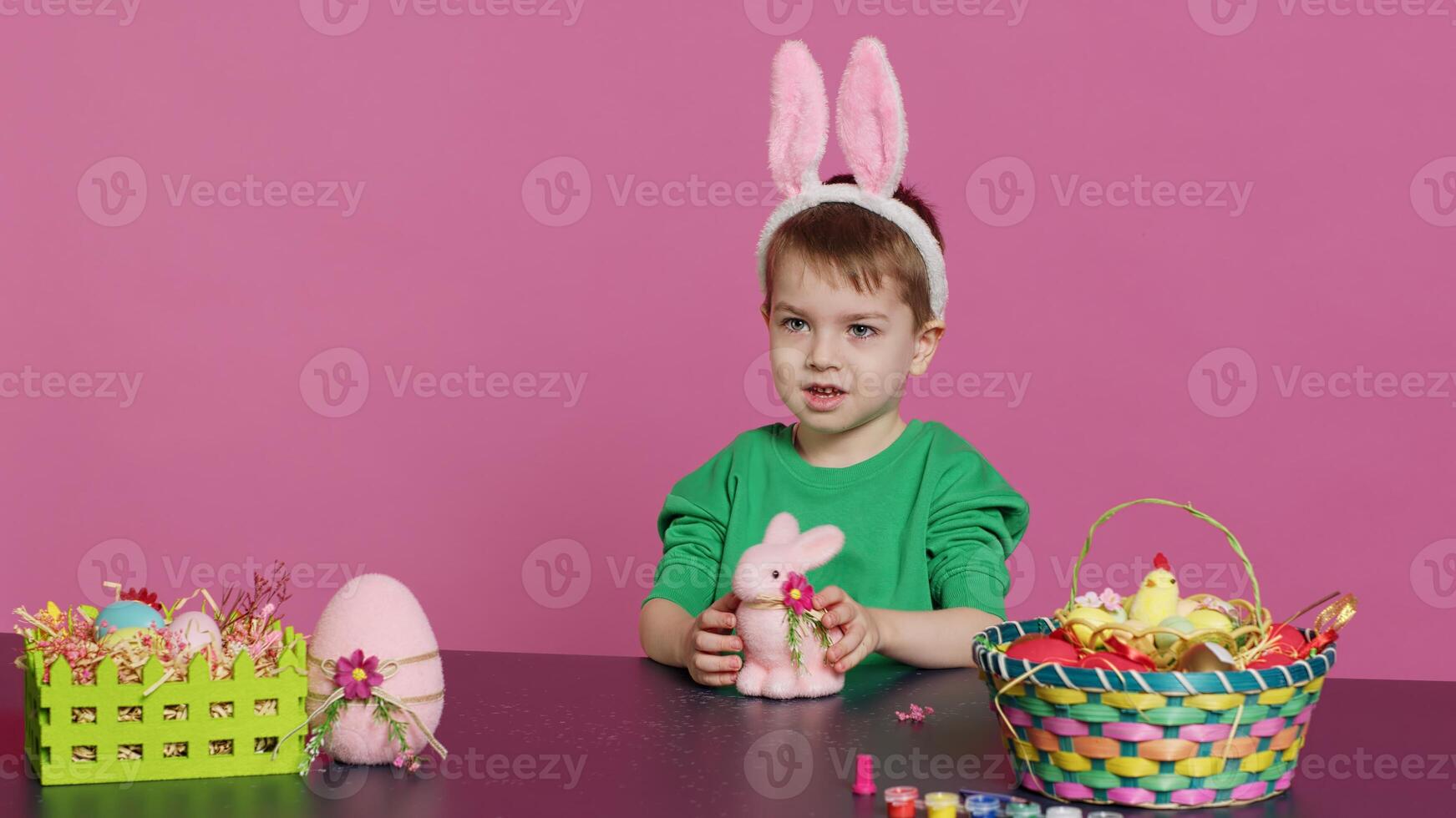 Adorable little child playing with a stuffed rabbit and a pink egg at table, having fun decorating festive easter ornaments in studio. Cheerful happy boy enjoying holiday preparations. Camera A. photo