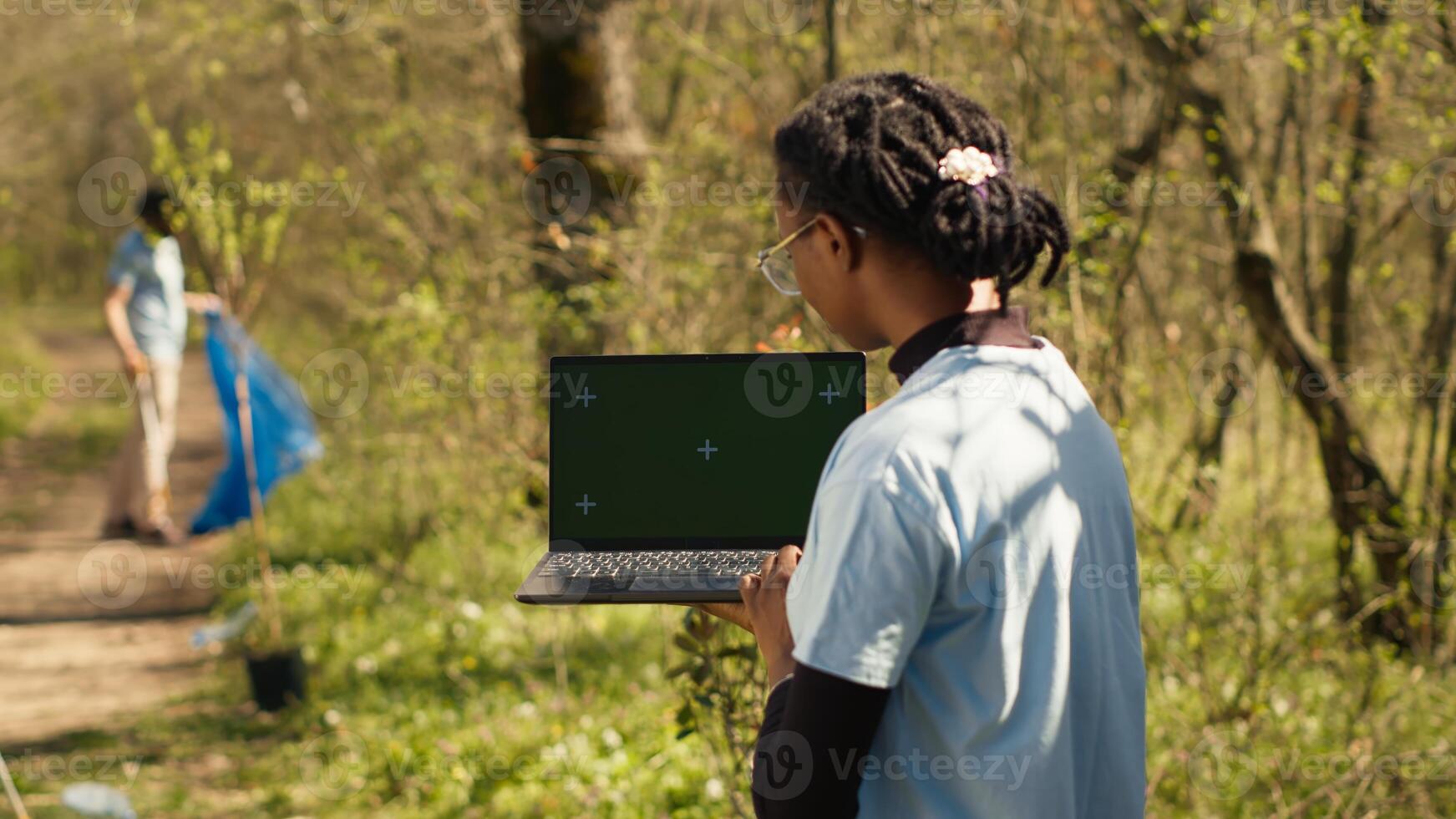 African american girl cleaning up forest and using isolated laptop display, taking part in a litter cleanup activity to protect the ecosystem and wildlife. Nature preservation concept. Camera A. photo