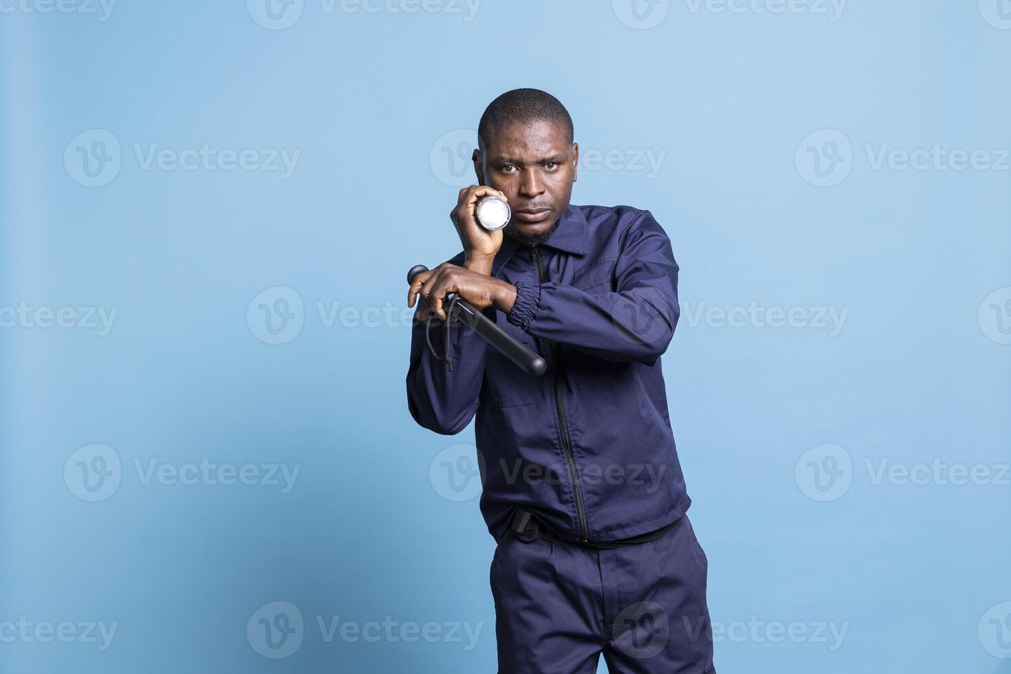 African american security agent on a patrol surveillance in the dark, using a flashlight against blue background. Bodyguard inspecting the perimeter, officer with a lantern for guidance. photo