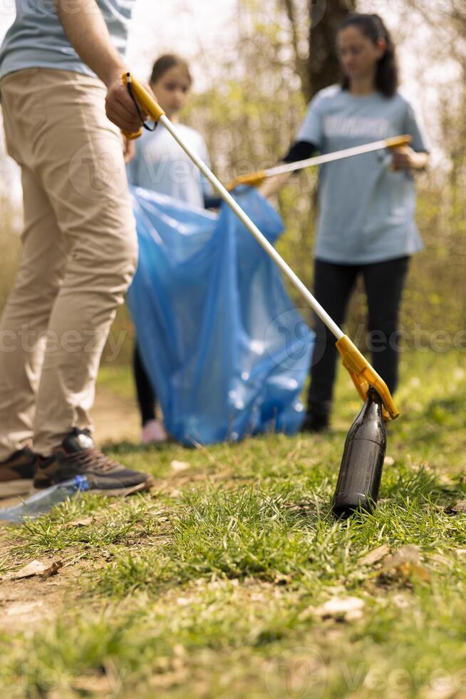 Environmentalists picking up trash from the natural forest environment by grabbing plastic and junk objects from the forest, collecting in a garbage bag. Using tools to pick up rubbish. photo