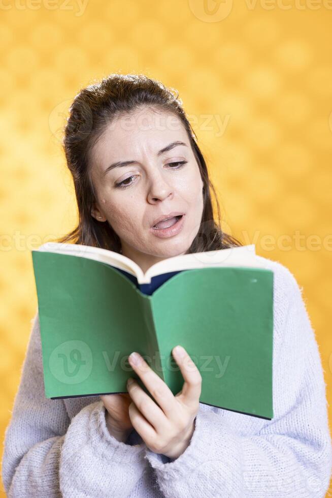 Shocked woman browsing through intriguing book pages, gasping, having fun, isolated over studio background. Surprised person reading stunning novel with plot leaving her with mouth open photo