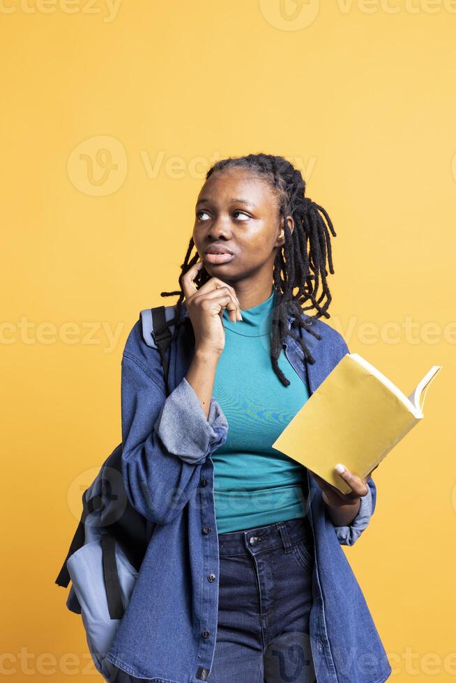 African american woman with book lost in thoughts, doing contemplation gesturing, isolated over studio background. Young girl holding novel, thinking about themes and characters after reading photo