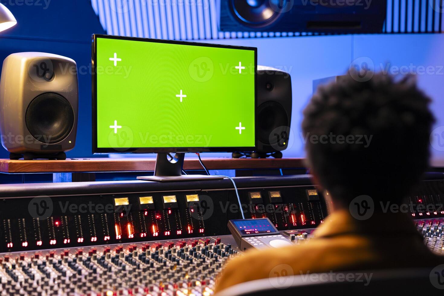 African american producer looks at greenscreen on monitor and mixing music with control desk panel board. Audio expert working in professional studio to adjust volume and add sound effects. photo