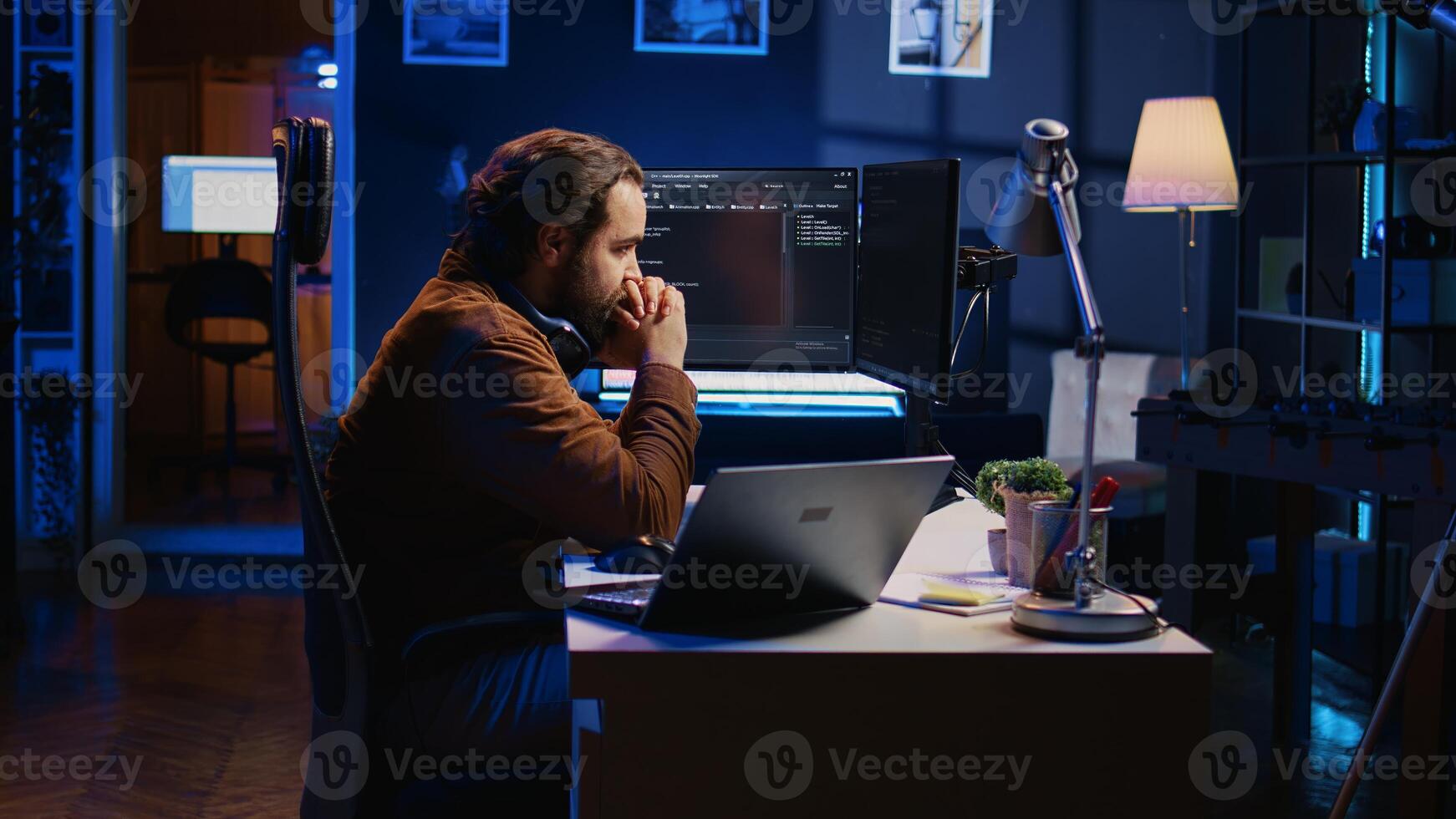 Technician focused on typing complex code in neon lit home office, developing software application. It specialist removing headphones to better concentrate on difficult task, camera B photo