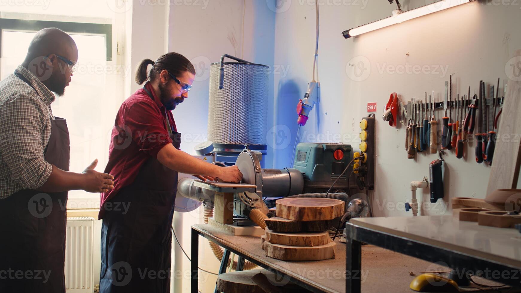 Man teaching african american trainee how to shape wooden bowl on disc sander in assembling shop. Worker learning from woodworking professional how to use grinding machine rotating disc, camera B photo