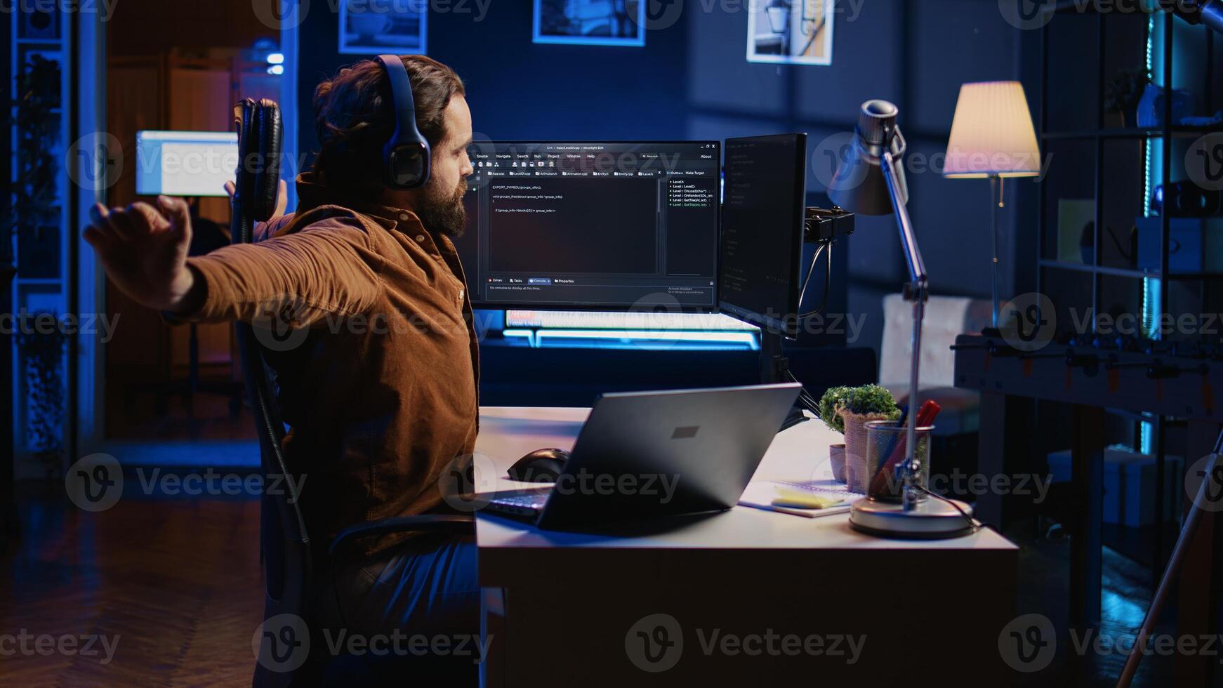 Software programmer taking break from coding, putting headphones on and stretching on desk chair during remote job shift. IT professional pausing tasks, listening music, loosening up, camera B photo
