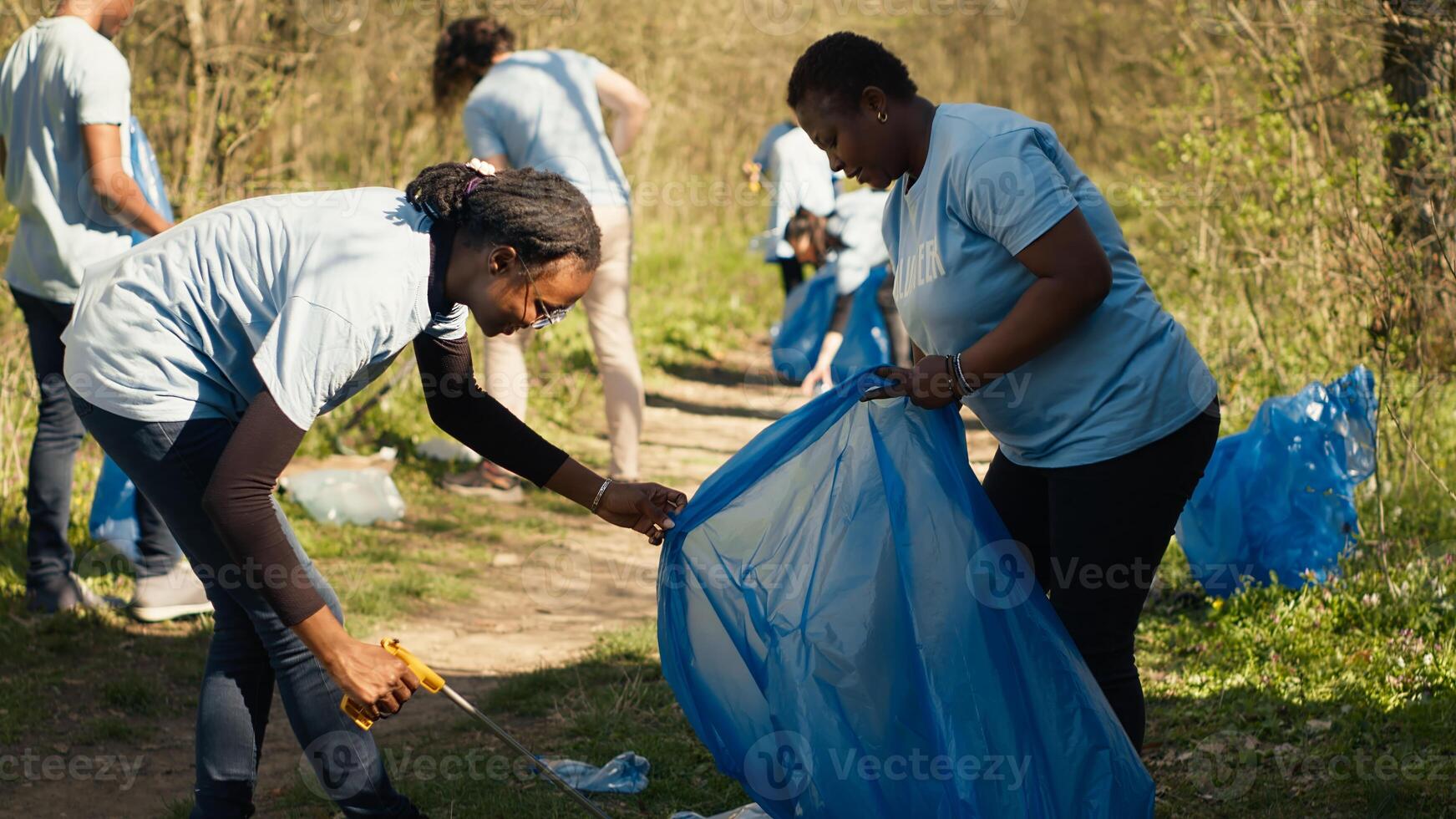 African american team of volunteers picking up trash from the ground, collecting rubbish and recycling in bags. Women cleaning the forest environment from plastic waste, illegal dumping. Camera A. photo