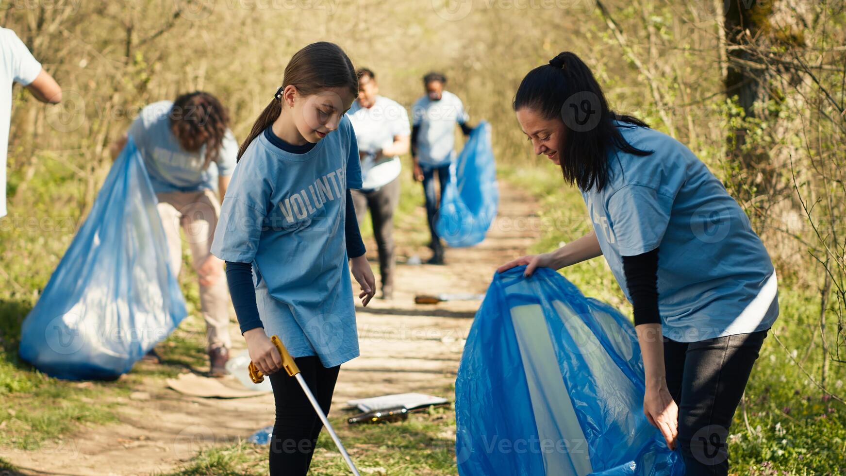 Mom and child collecting rubbish from the forest area using a long claw tool and garbage disposal bags. Little girl picking up trash and plastic waste with her mother, ecological justice. Camera A. photo