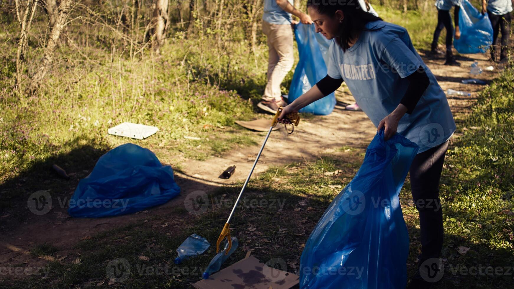 Environmental activist picking up trash with a claw tool and recycling, protecting the natural forest habitat and doing litter cleanup. Woman doing voluntary work to stop pollution. Camera B. photo