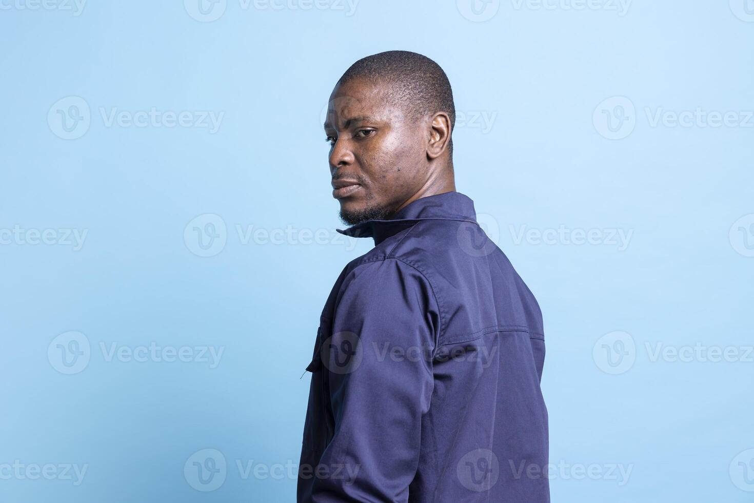 Portrait of african american security agent posing in front of camera, looking over his shoulder against blue background. Male bodyguard dressed in uniform, detecting any suspicious behavior. photo