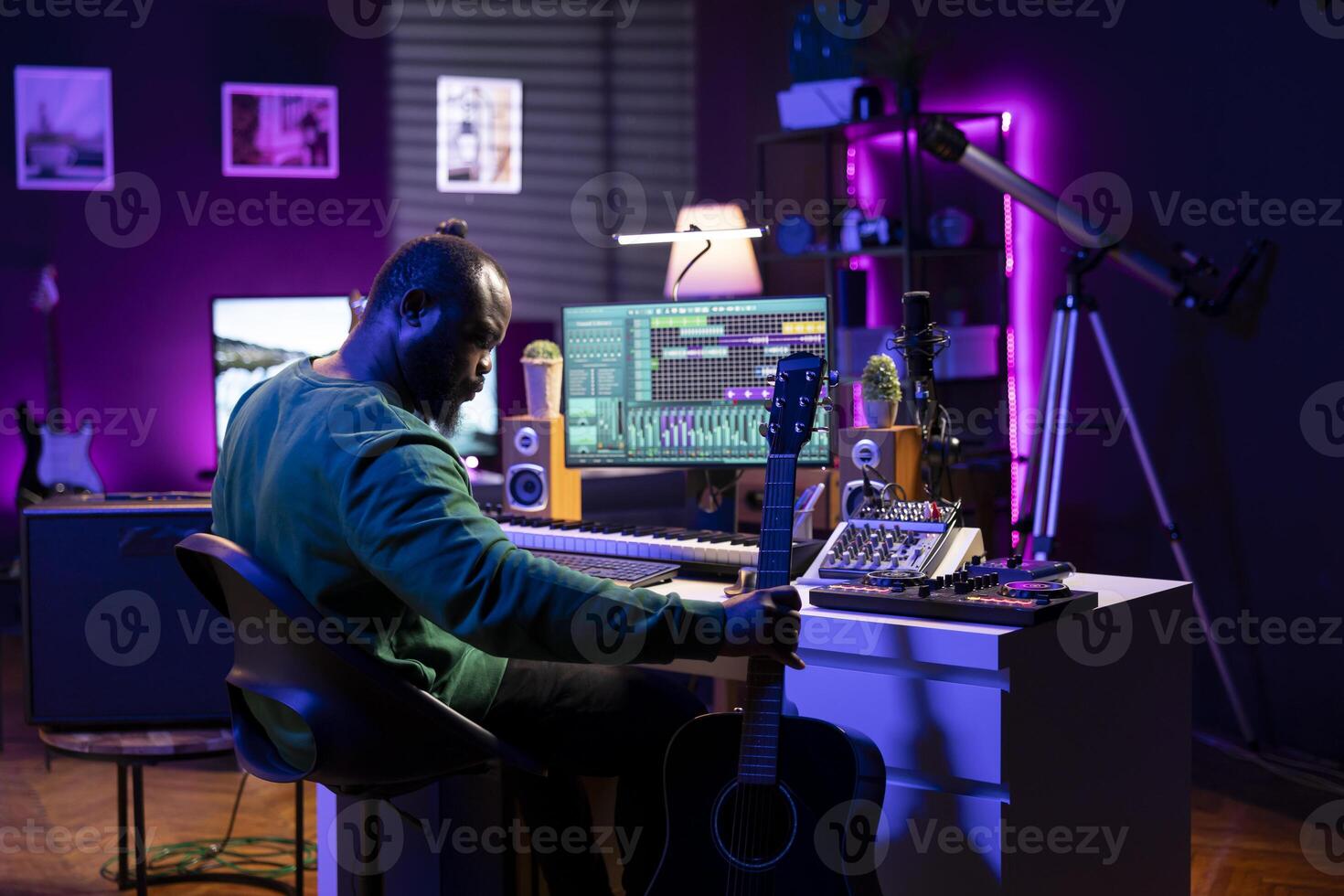 Producer composing new music on acoustic instrument in home studio, using a microphone to sing the chords. Musician songwriter works with mixing console and mixing software, guitarist producing tunes. photo