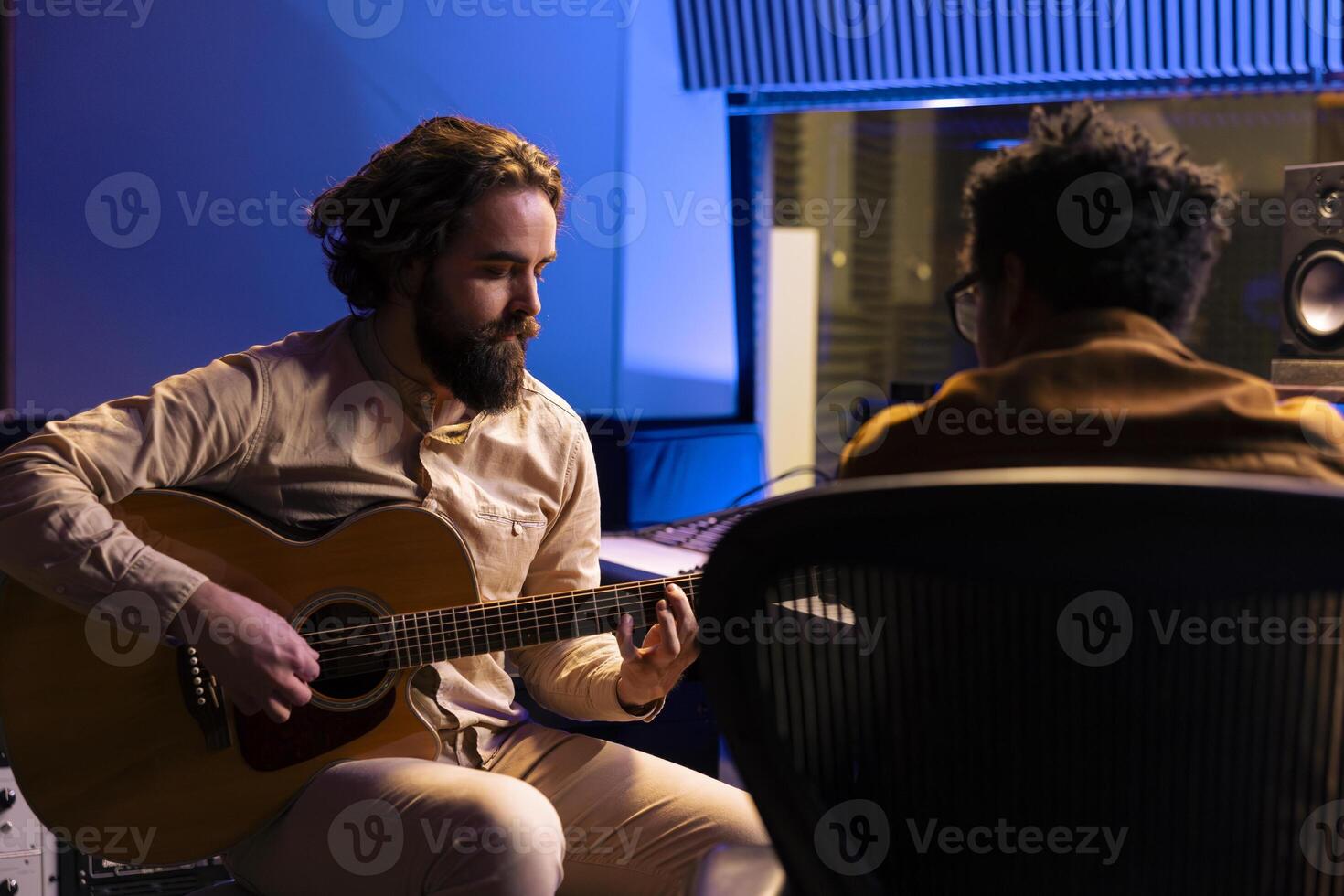 Singer playing electro acoustic guitar to record his tunes and compose a hit song, collaborating with audio technician. Young skilled artist performing music in professional studio, post production. photo