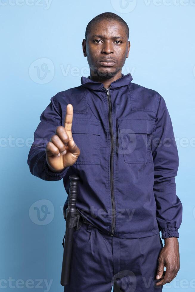 Serious security warden showing his authority with a restriction sign, limiting the access for people by raising a finger up in studio. Patrol agent in uniform does stop symbol as interdiction. photo