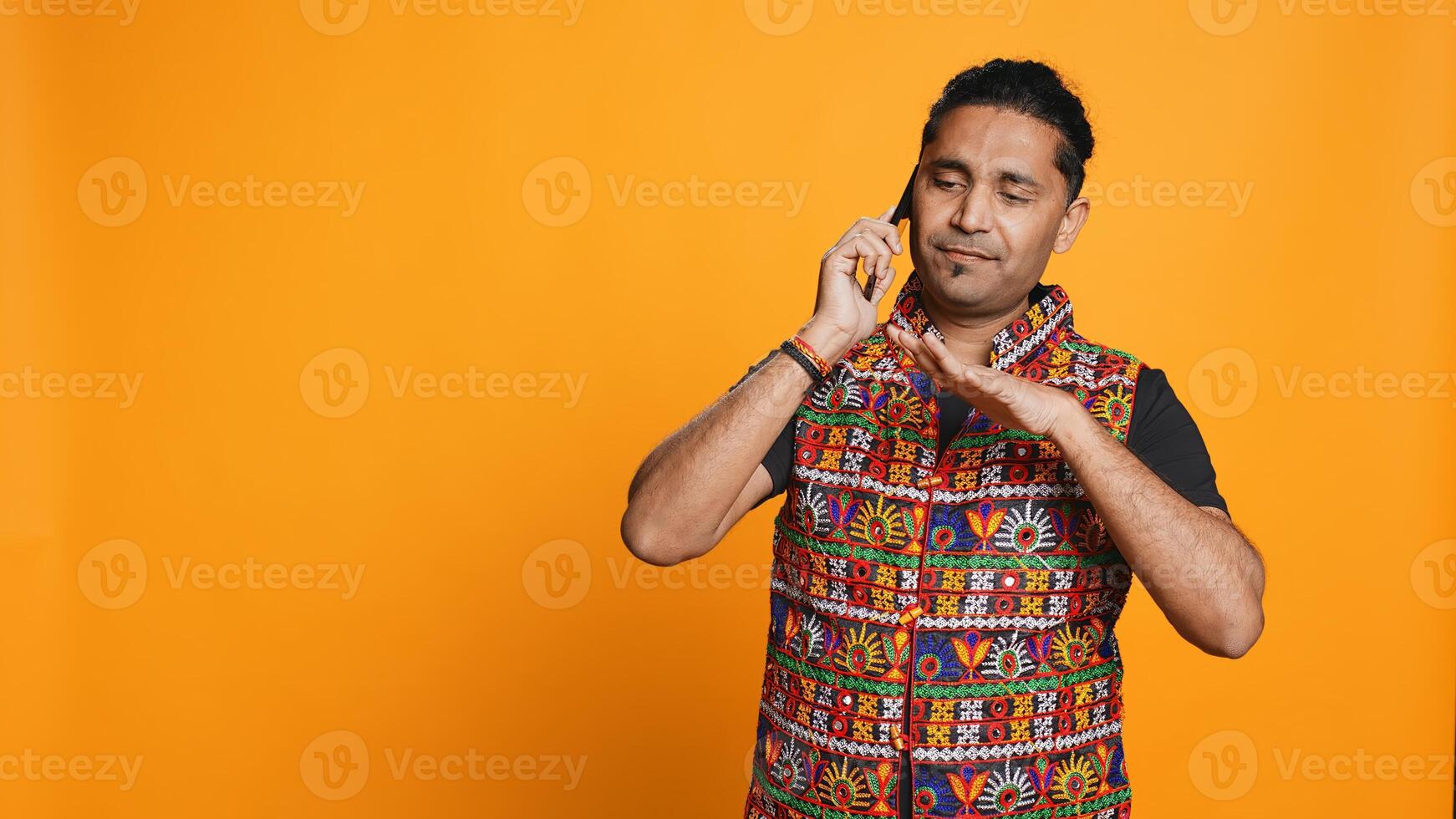 Happy man in traditional clothing chatting with best friend over telephone call. Upbeat person with colorful attire communicating with mate using phone, studio background, camera B photo