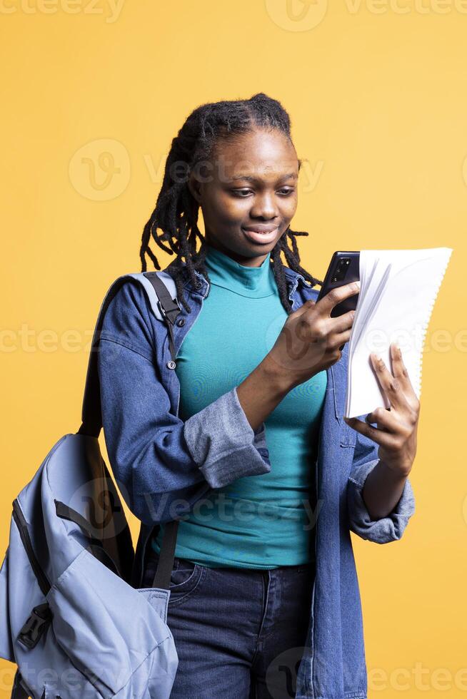 BIPOC young girl taking photos of school homework to send to colleague, isolated over studio background. Student photographing papers with information needed for university courses using phone