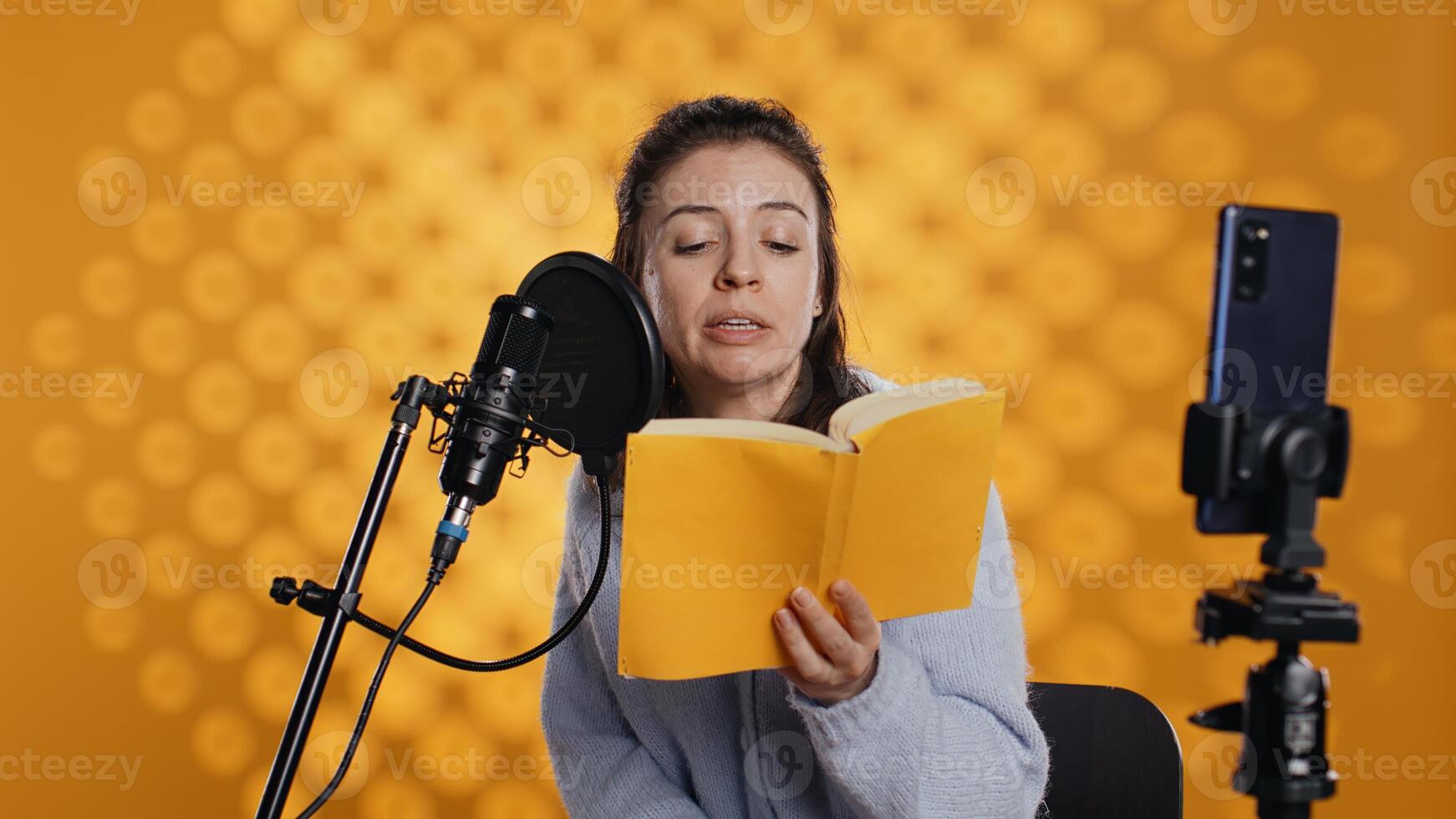 Influencer filming reading book with smartphone on tripod, creating audiobook, studio background. Woman vlogging with mobile phone while producing digital recording on novel using microphone, camera B photo