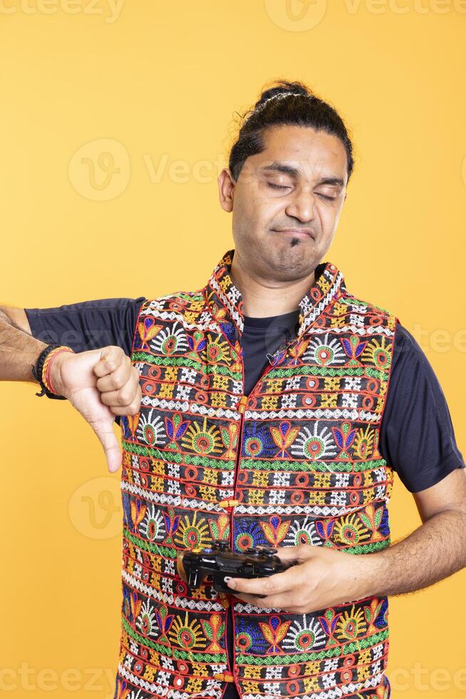Portrait of annoyed man showing thumbs down sign holding gamepad, disapproving of videogames. Disgusted person protesting against gaming, doing rejection hand gesture, isolated over studio backdrop photo