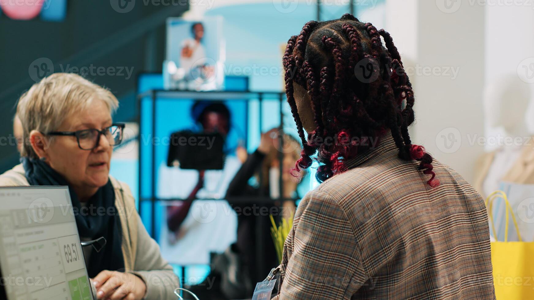 Elderly woman choosing clothing items to by from showroom, approaching cash register to pay for products. African american mall employee assisting senior customer at checkout. Camera A. photo