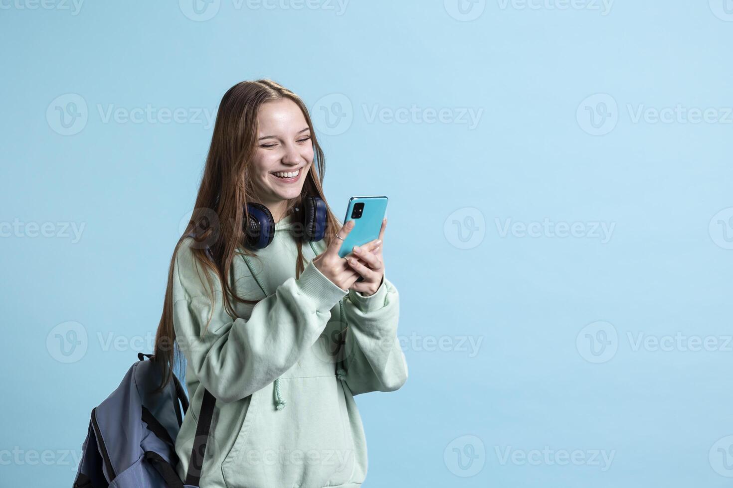 Radiant teenager using smartphone, laughing at amusing social media posts on feed. Joyful person browsing internet websites on phone, amused by funny memes, studio backdrop photo