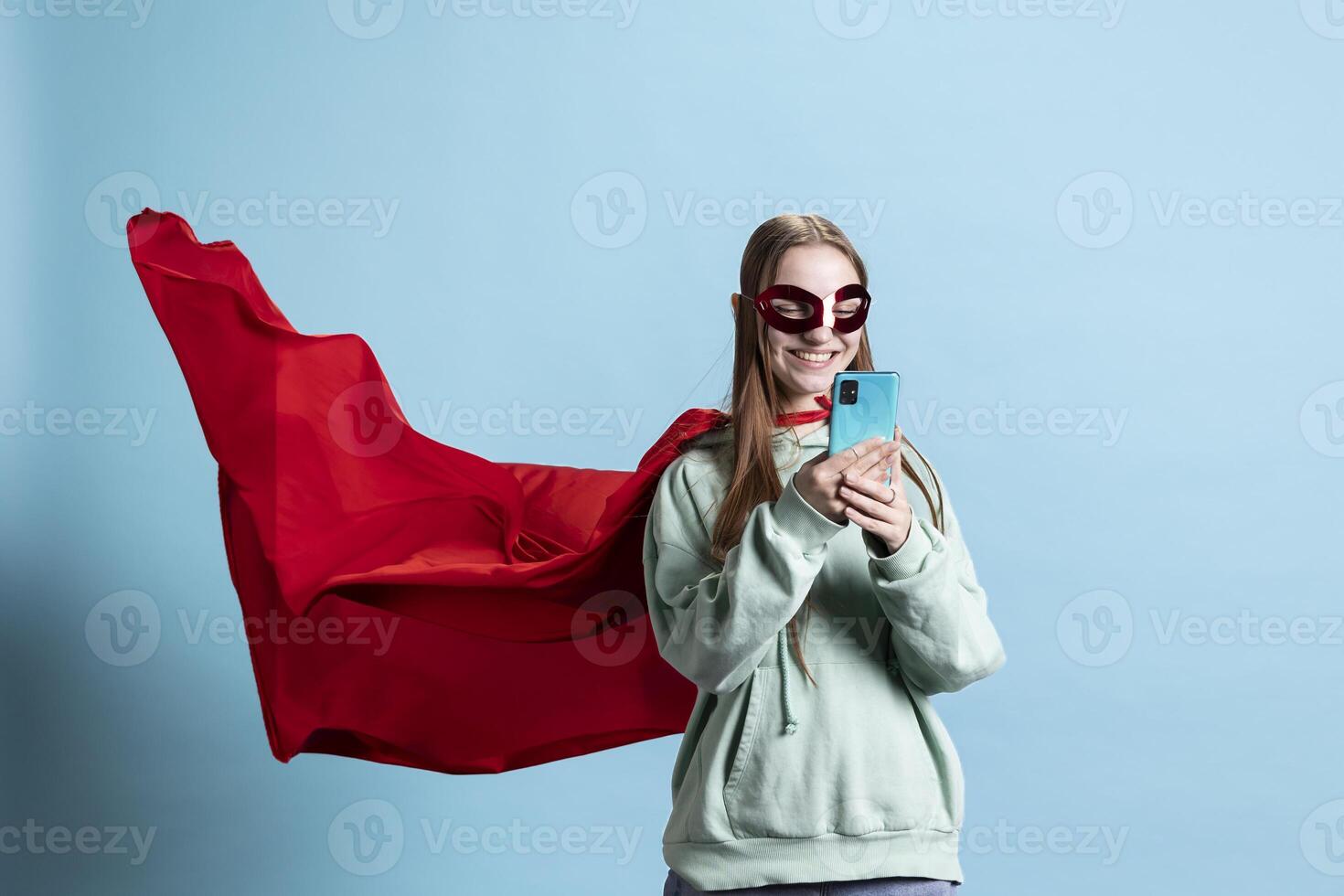 Smiling girl with mask portraying superhero, texting friends on mobile phone, studio background. Happy woman posing as hero in costume with mantle sending messages on smartphone photo
