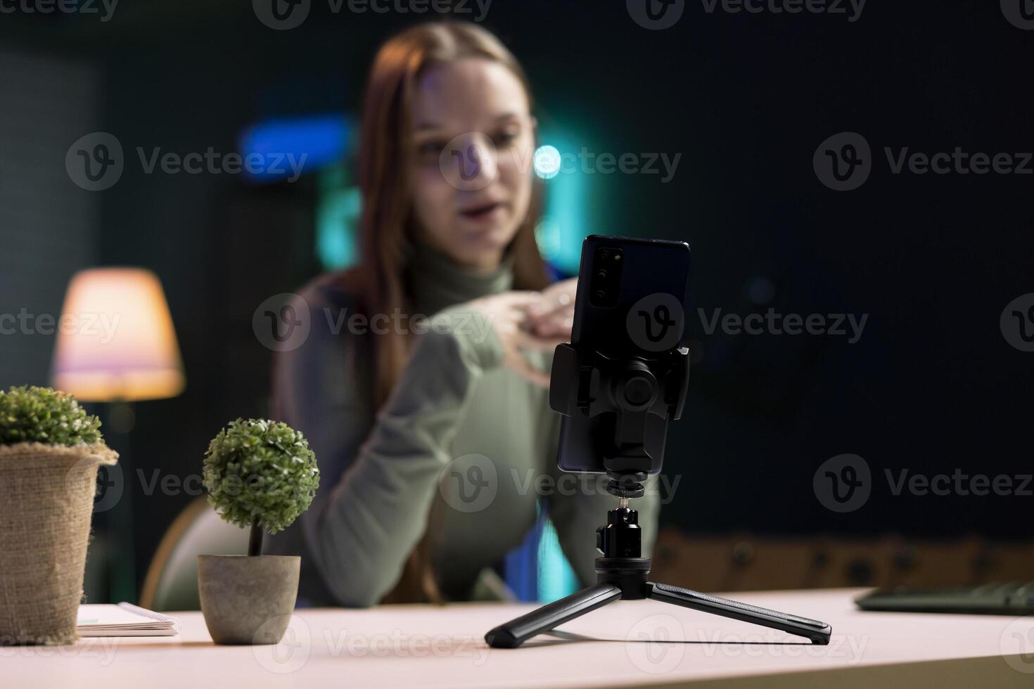 Focus on cellphone on tripod used by young entertainer in blurry background to film in living room used as professional studio. Close up of mobile phone used by gen Z person to record vlog photo