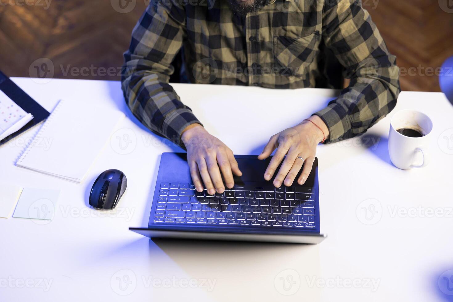 Person sitting at table surrounded by papers, cup of coffee and electronic device. Top view image of male individual typing and surfing the net on his laptop. photo