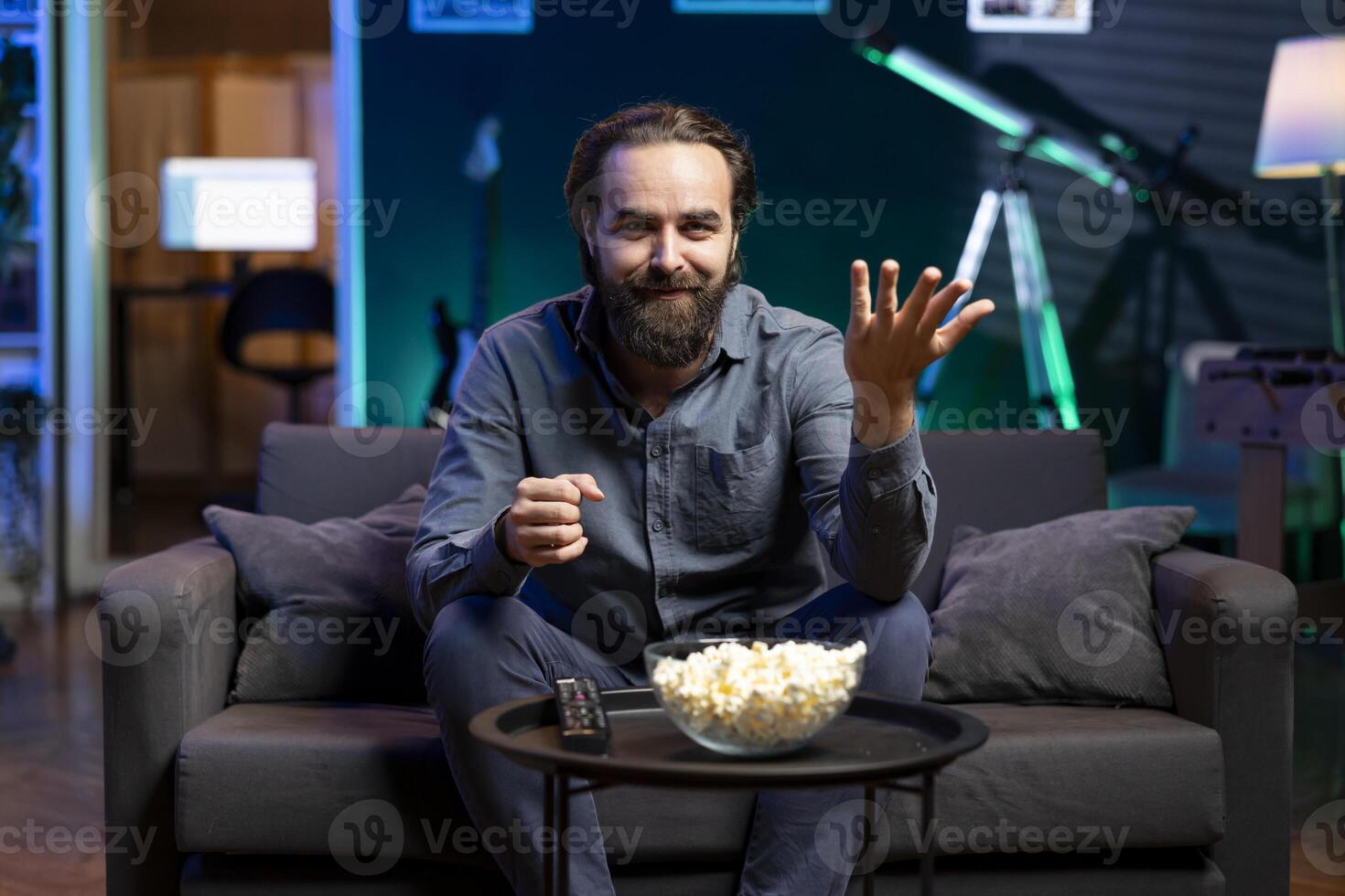 Joyous man sitting on couch laughing at funny comedy movie while eating from bowl of popcorn. Upbeat person enjoying leisure time in apartment, amused by hilarious humor in film scene photo