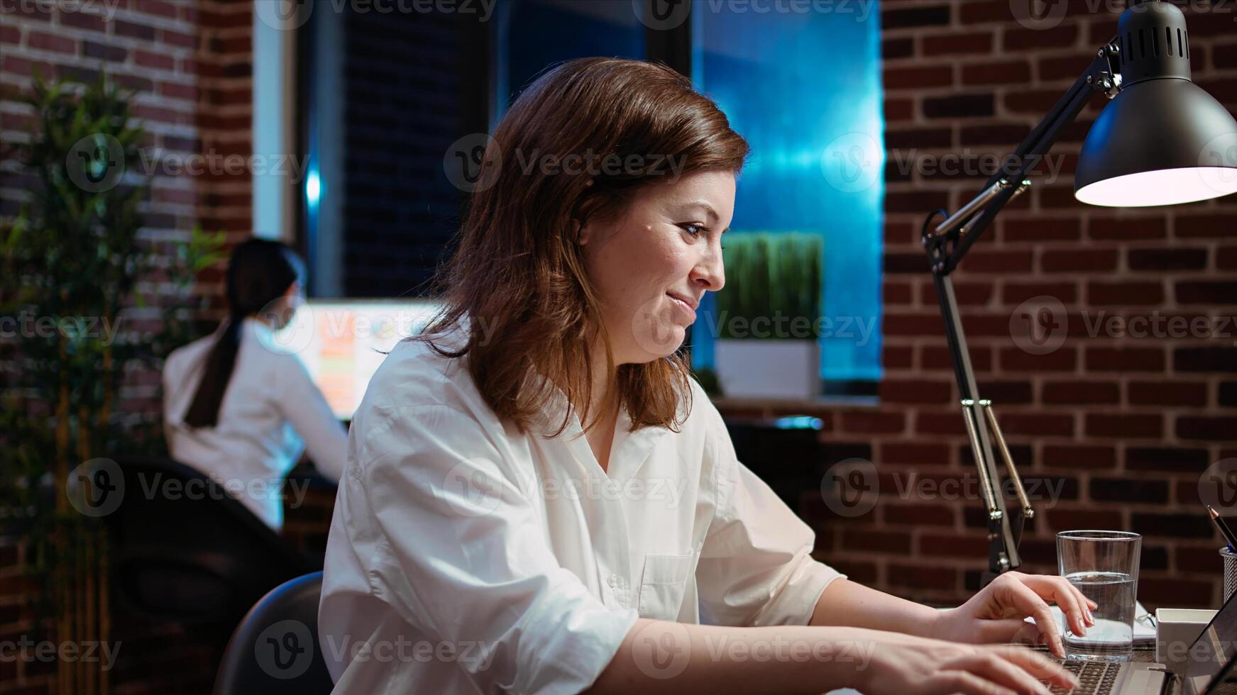 Close up of smiling businesswoman working for company project, doing tasks in brick wall office overnight. Cheerful employee inputting data on laptop late at night, zoom in shot photo