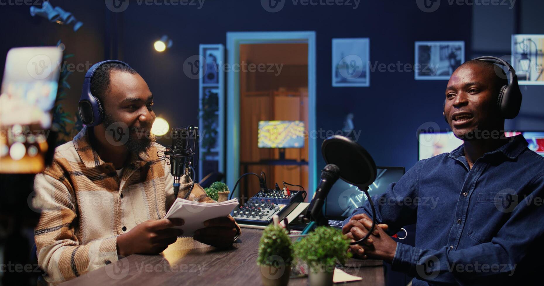 Men laughing during comedy focused podcast filmed with smartphone on tripod. African american guest amused by talking show host telling jokes, recording with mobile phone photo