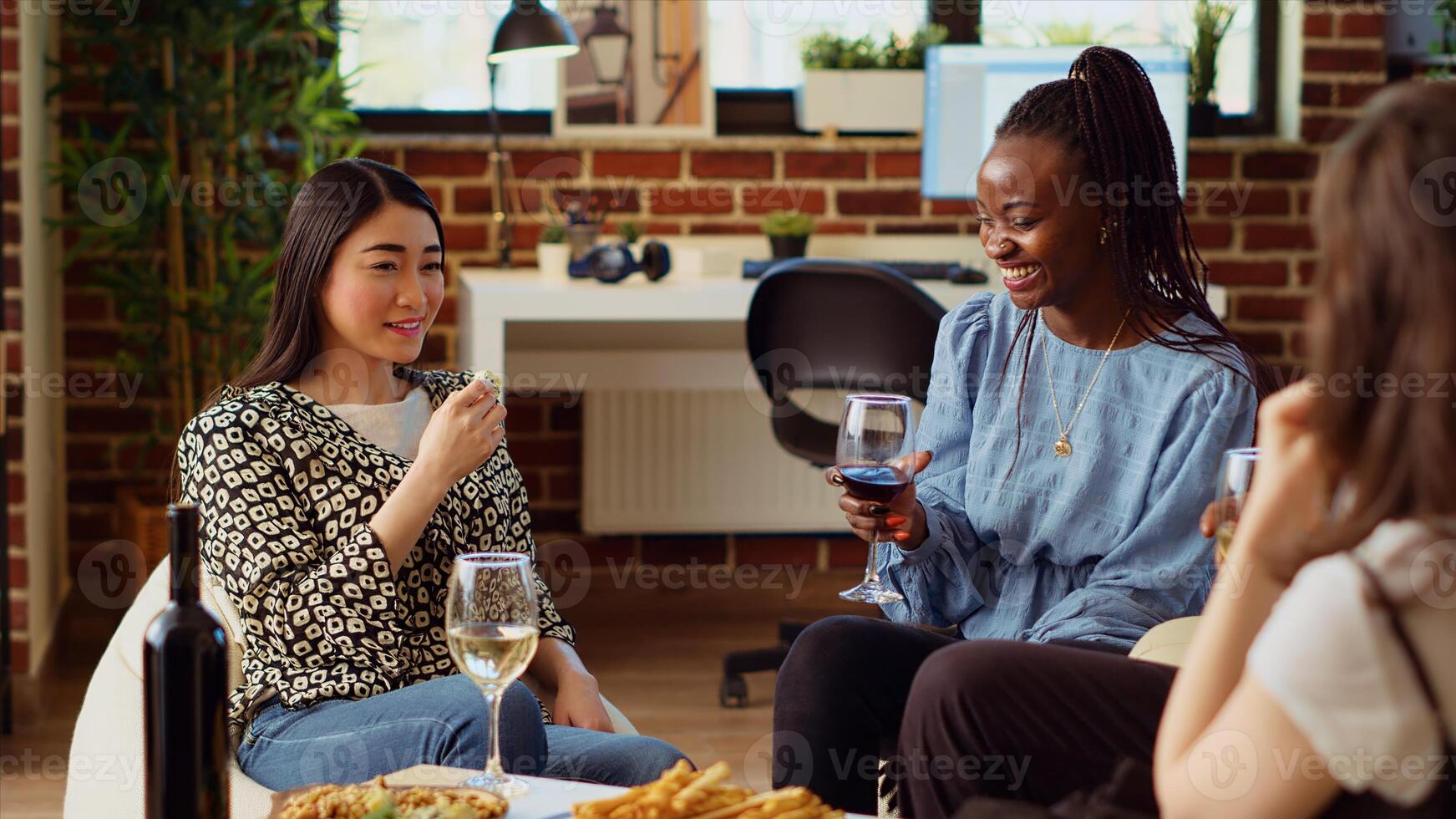Asian woman chatting with multiracial apartment party guests while eating appetizers. Host enjoying gourmet cheese from charcuterie platter while communicating with friends photo