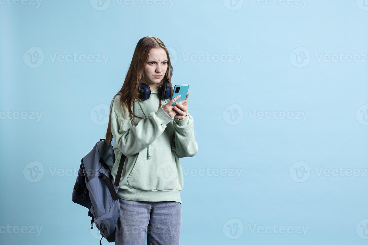 Teenager frowning while reading messages on phone, feeling shocked after receiving bad news. Woman feeling confused by weird SMS on smartphone, isolated over studio background photo