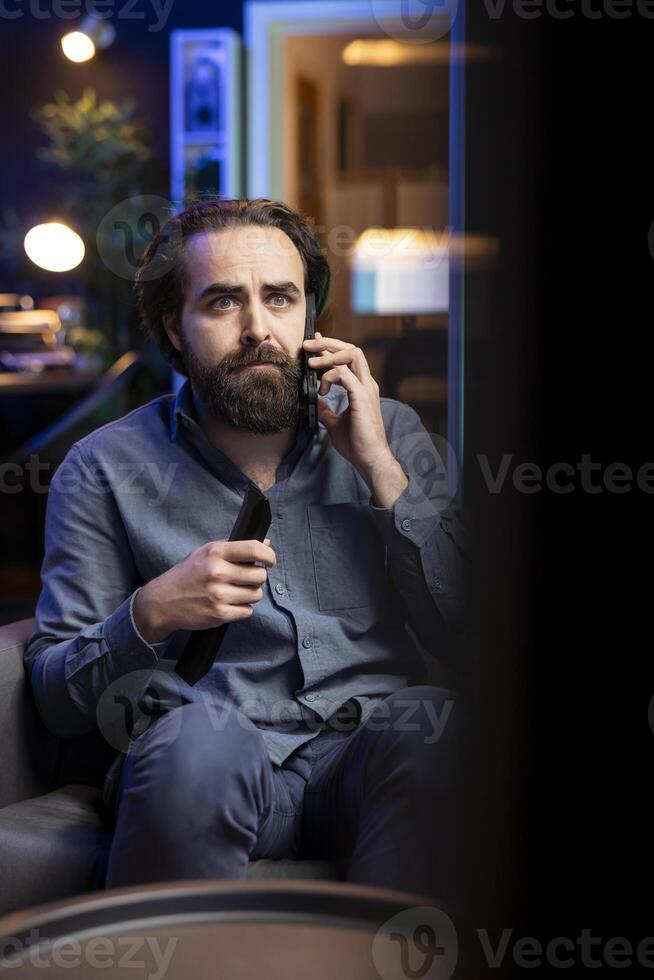 Sad man on couch watching heartbreaking drama movie on streaming service while talking on phone. Person distressed by TV show favorite character dying, discussing with friend on smartphone photo