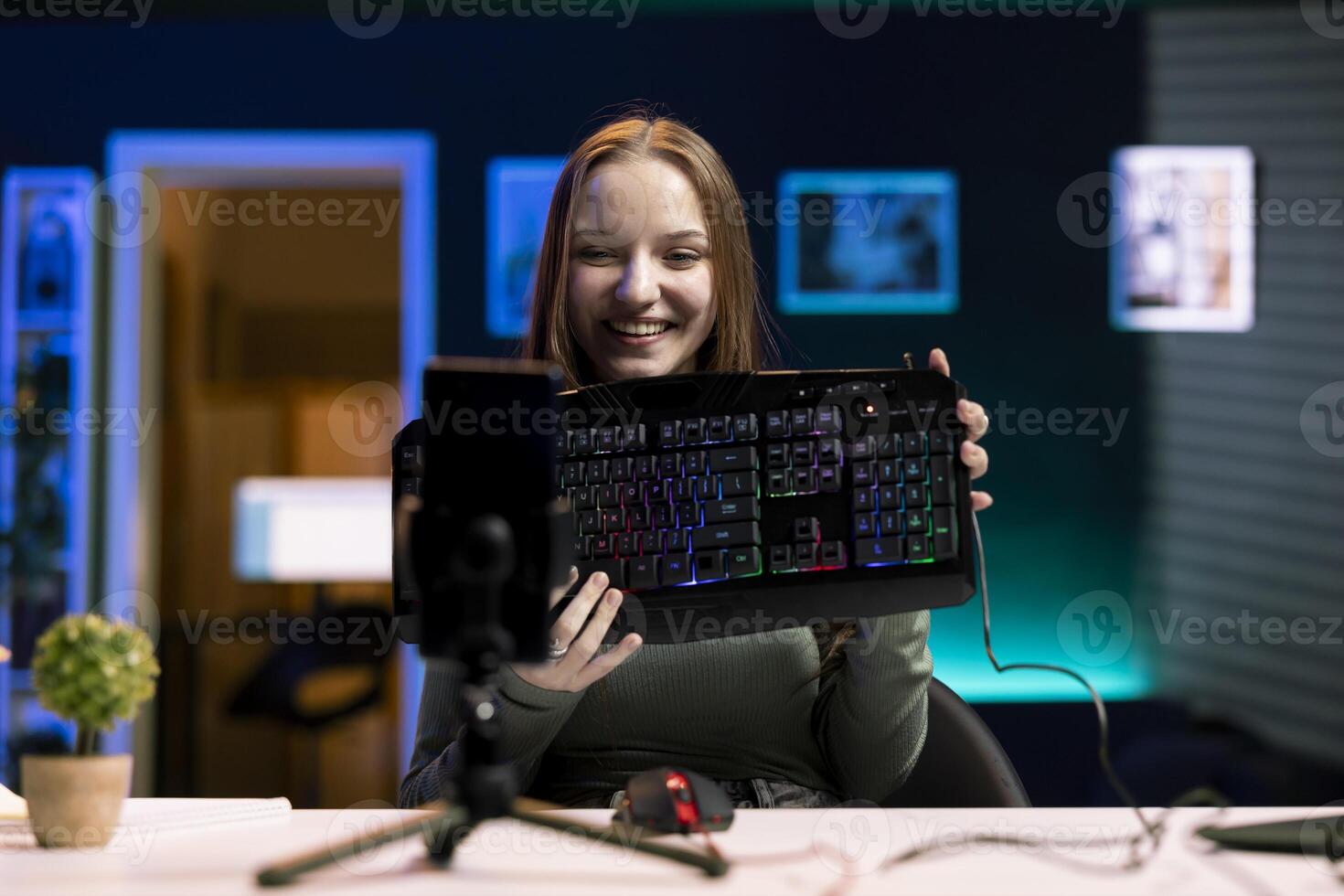 Joyous girl reviews keyboard from sponsor, doing review for online streaming channel using mobile phone. Smiling media star excited to review computer peripherals brand, filming with smartphone photo