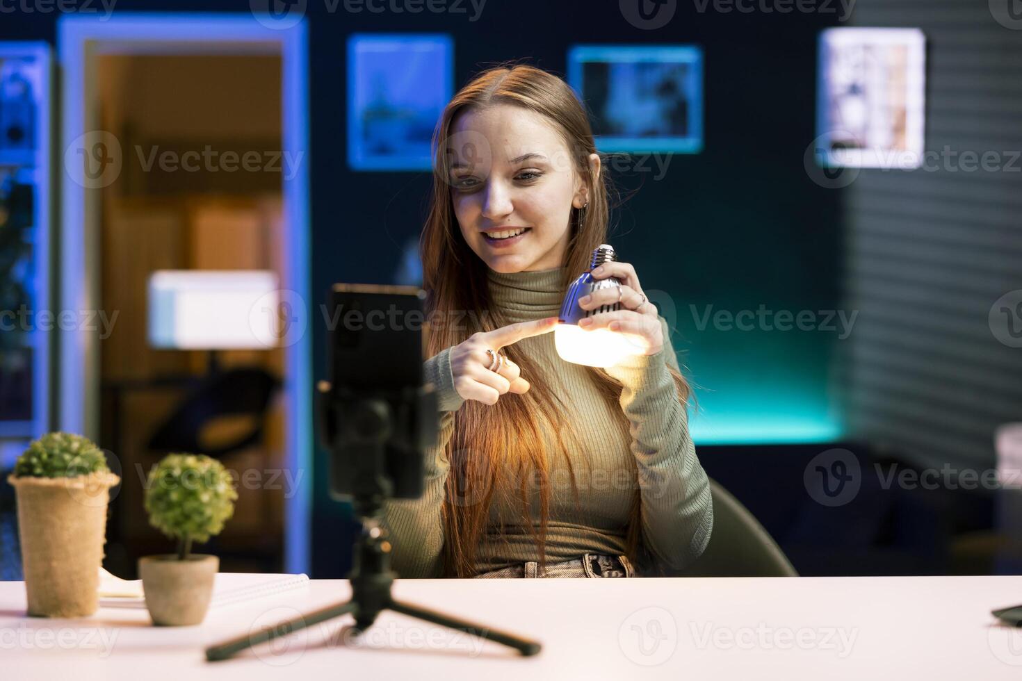 Happy girl doing house improvement vlog, filming newly launched smart light bulb with phone. Smiling entertainer greeting fanbase, presenting WIFI home automation, recording with smartphone photo