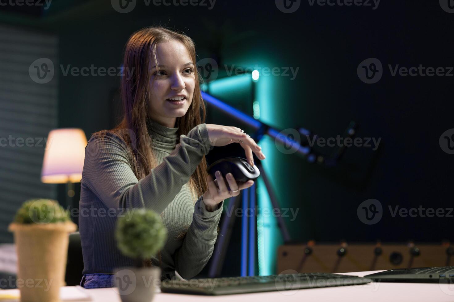 Girl filming for online channel in dimly lit home studio, presenting wireless mouse to audience. Teenage content creator showing audience computer peripheral device photo