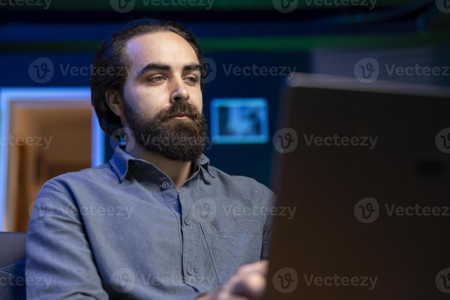 Man on couch working from home in apartment solving business related tasks on laptop. Teleworking company manager on couch in neon lit living room sending emails on notebook photo