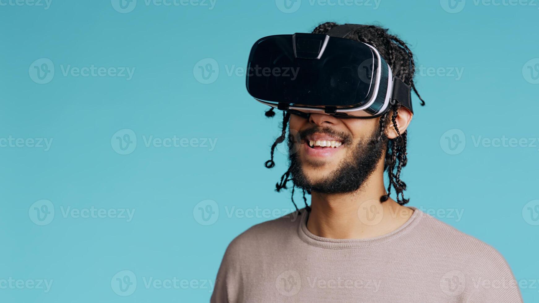 Middle Eastern man wearing virtual reality headset, doing swiping gestures. BIPOC person using high tech futuristic modern VR gadget, isolated over blue studio background, camera B photo