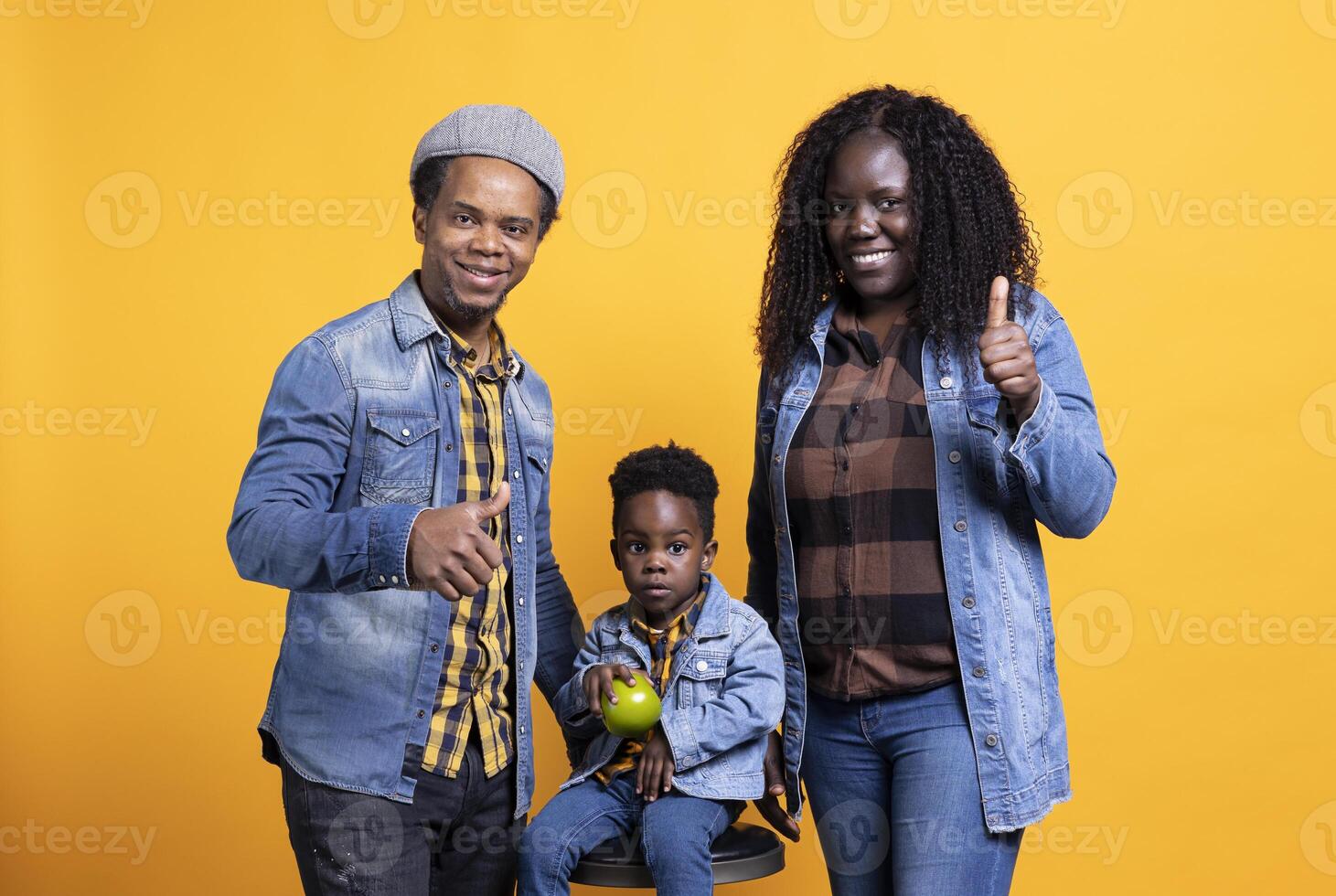 African american mom and dad giving like symbol in front of camera, posing with their adorable son sitting on the chair. Joyful young parents showing thumbs up sign, approving in studio. photo