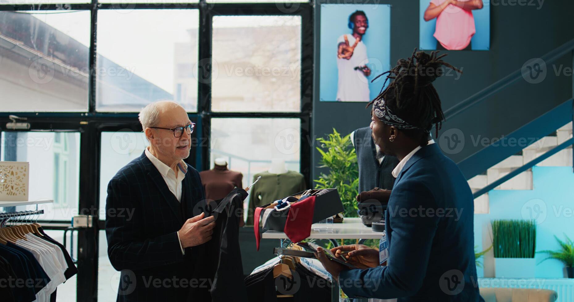 Regular elderly client asking store worker for assistance with clothing items, looking for a shirt to go with his suit jacket. African american clerk managing inventory and helping shopper. Camera B. photo