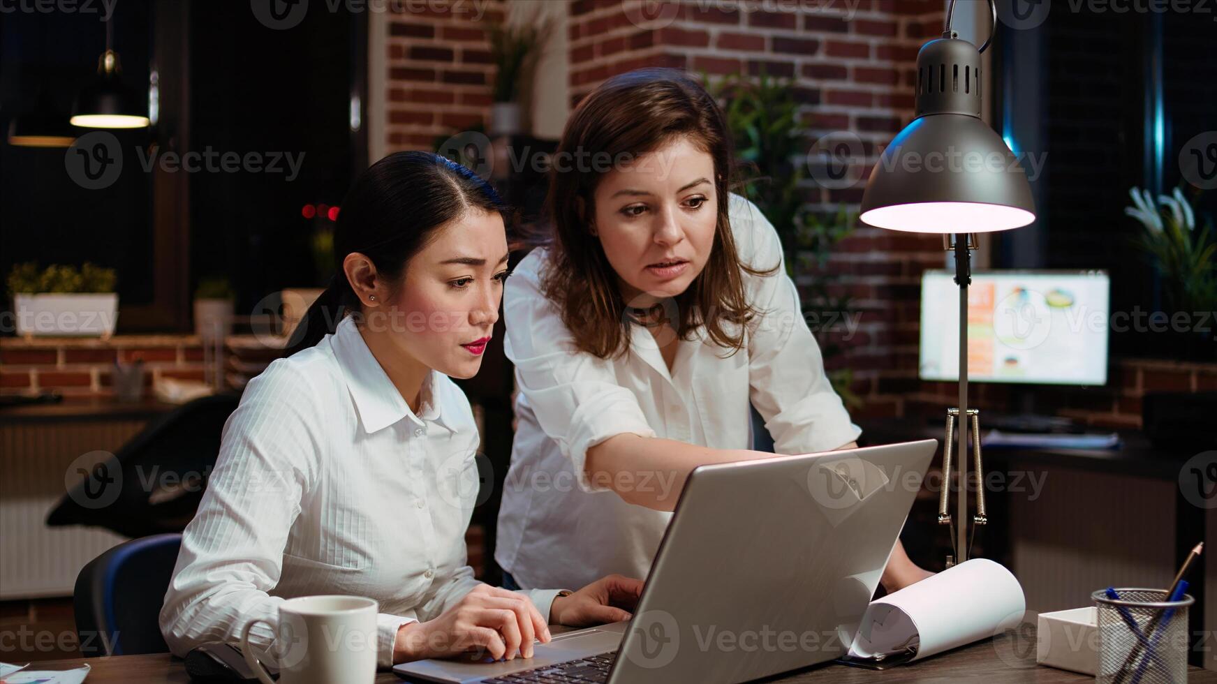 Asian businesswoman and colleague working together doing computer tasks for team project in office overnight. Coworkers looking over accounting figures on laptop screen late at night photo