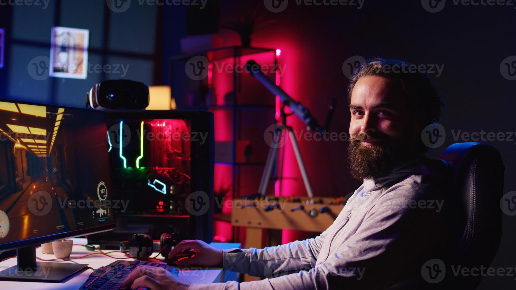 Portrait of smiling man in dark living room playing engaging games on gaming PC at computer desk, chilling after work. Happy gamer contending against foes in online multiplayer shooter photo