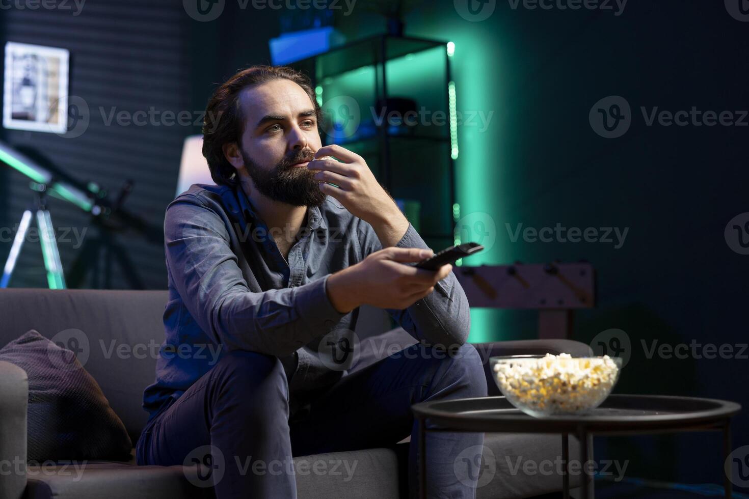 Man in neon lit room on couch eating popcorn while browsing though movies available on streaming service, enjoying time off from work. Viewer consuming snacks at home, holding remote control photo