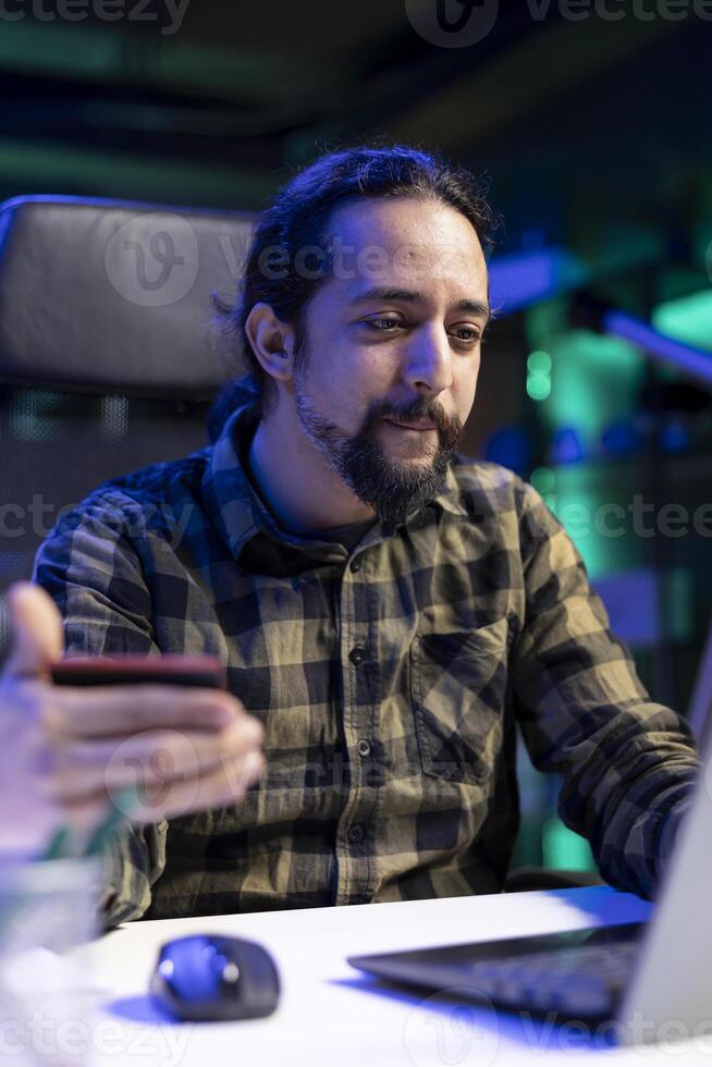 Male individual with credit card, purchasing in an online store making wireless payments at home. Young bearded man using laptop to buy and pay for products. Electronic shopping concept. photo