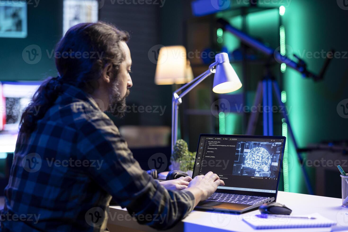 Youthful man using machine learning to manage a complex system on his laptop. Male individual sitting at his desk and typing code to create an online neural network database. photo