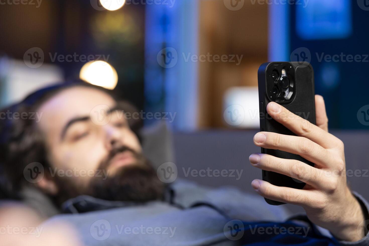 Focus on smartphone with man in blurry background lounged on couch texting friend inviting him to watch movie on streaming service. Close up on phone used by person at home discussing with mate photo