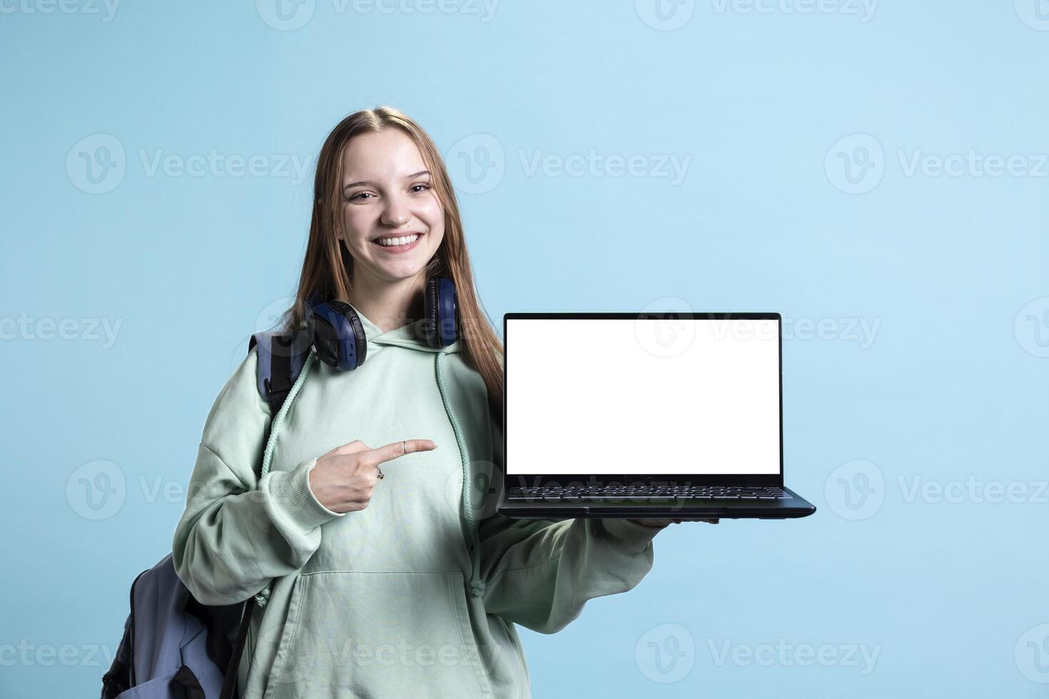 Happy young girl laughing while presenting laptop with isolated screen display, studio background. Upbeat teenager has fun during promotion, pointing finger towards mockup notebook photo