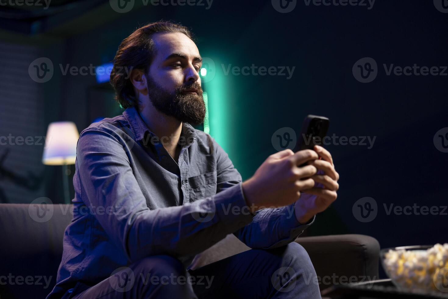 Person in living room binging series on streaming service, texting friend on mobile phone. Man with bowl of popcorn in front watching shows on television and chatting with mate on smartphone photo