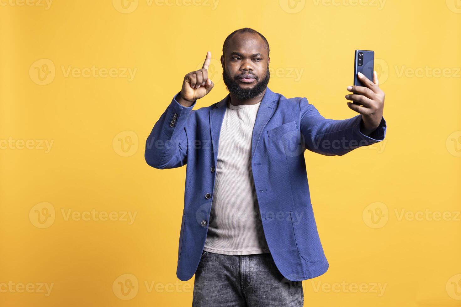 Man using smartphone to take selfies, pointing finger up. BIPOC person taking photos using phone selfie camera, doing hand gesturing, isolated over yellow studio background