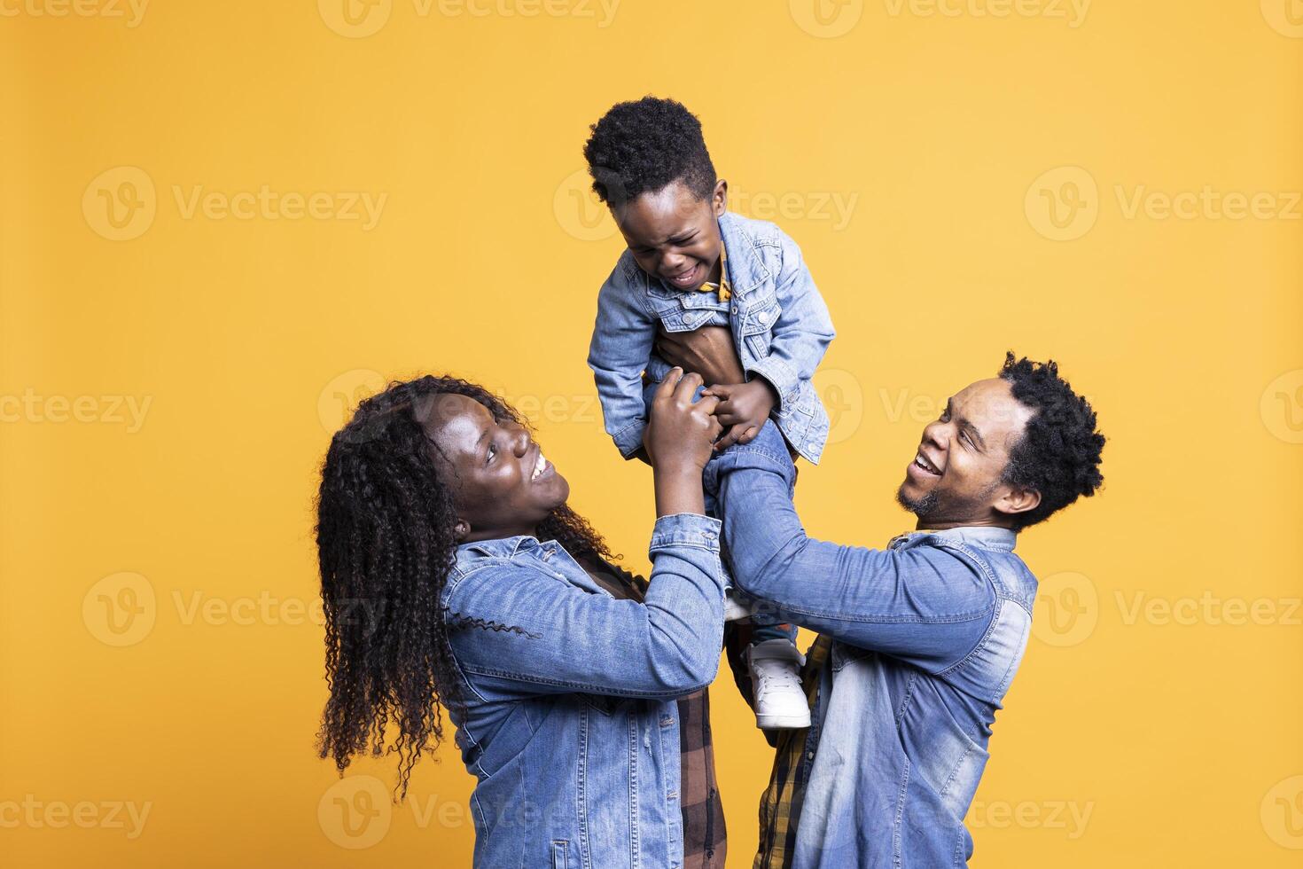 Loving african american parents lifting their cute small son in studio, posing in front of the camera. Proud happy couple bonding with their adorable toddler against yellow background. photo